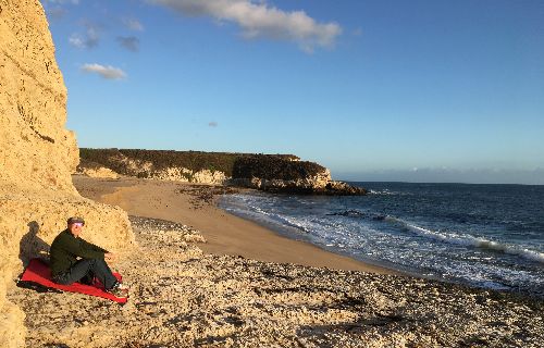 Professor Sparke sits on a red blanket at the left foreground, the beach with waves are breaking to the right, a costal bluff is in the distance, a few clouds in a blue sky